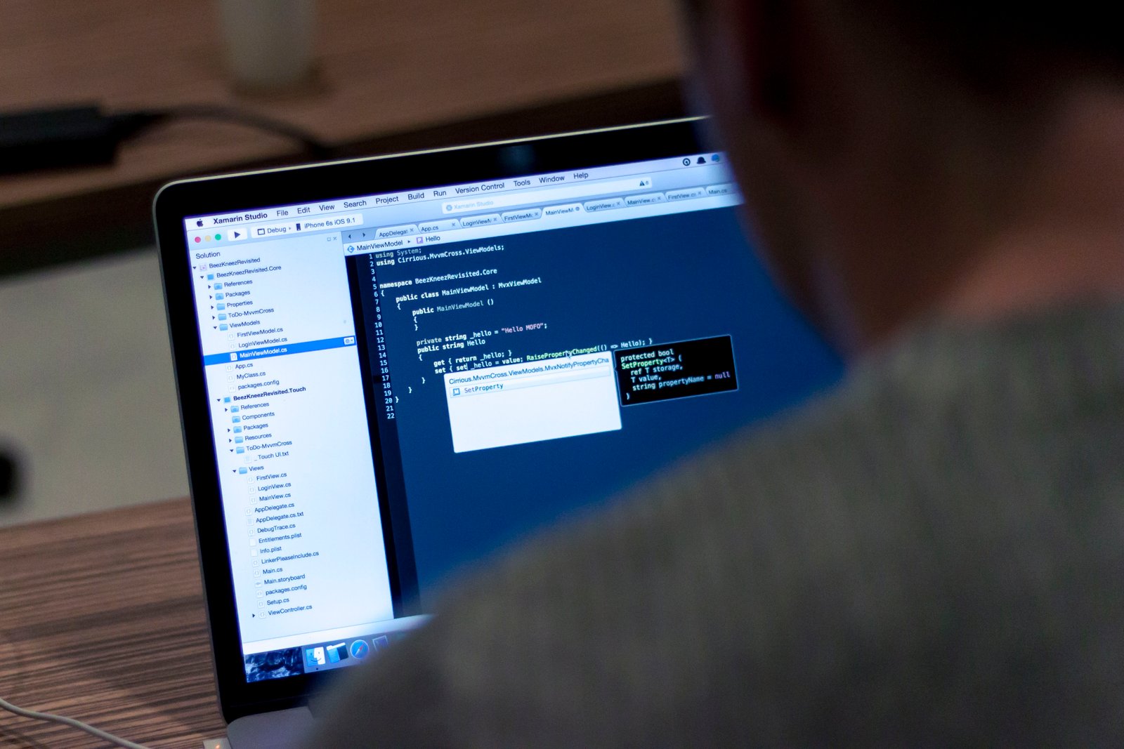 Over-the-shoulder view of a developer at a laptop reading C# source code in an IDE, the editor window glowing blue against a dark room.