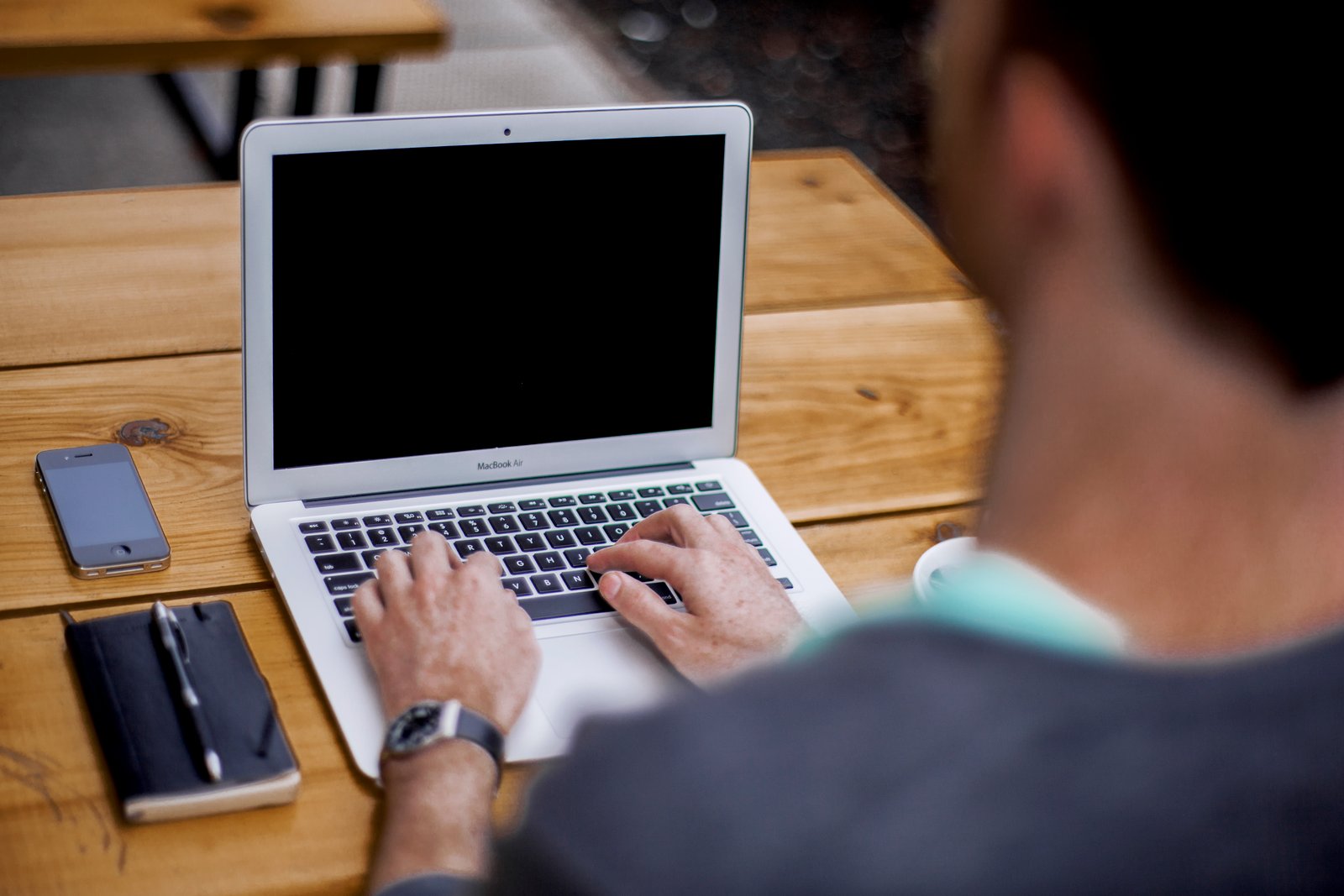 A man typing on a MacBook Air laptop at a wooden desk, with a notebook and smartphone nearby