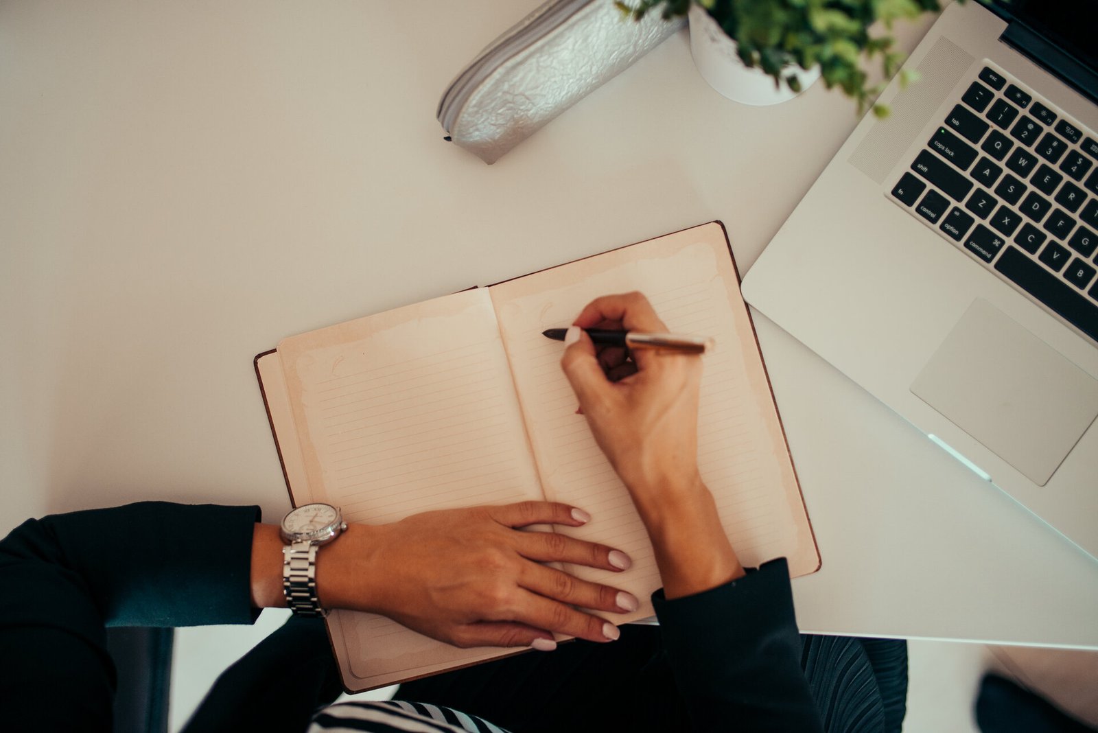 Top-down view of a person writing in an open notebook with a pen, with a laptop and small plant on a sleek white desk.