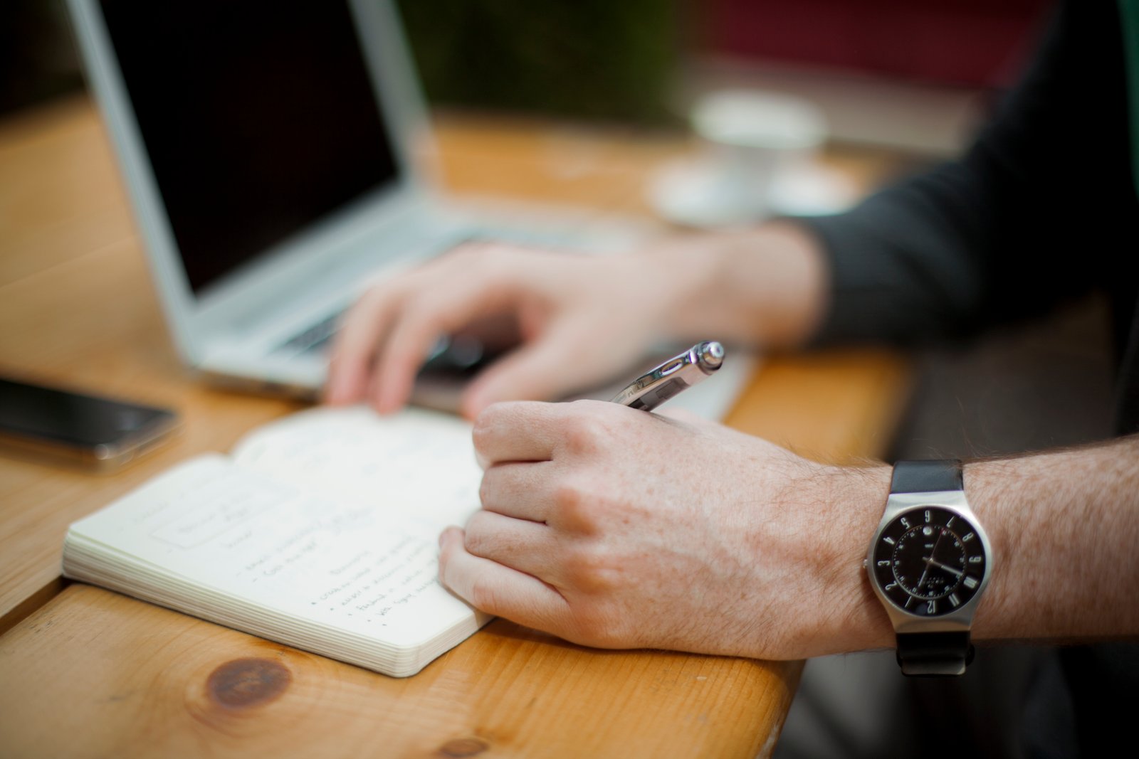 Two hands writing in a notebook beside a laptop on a wooden desk, one wrist wearing a watch, the other holding a pen.