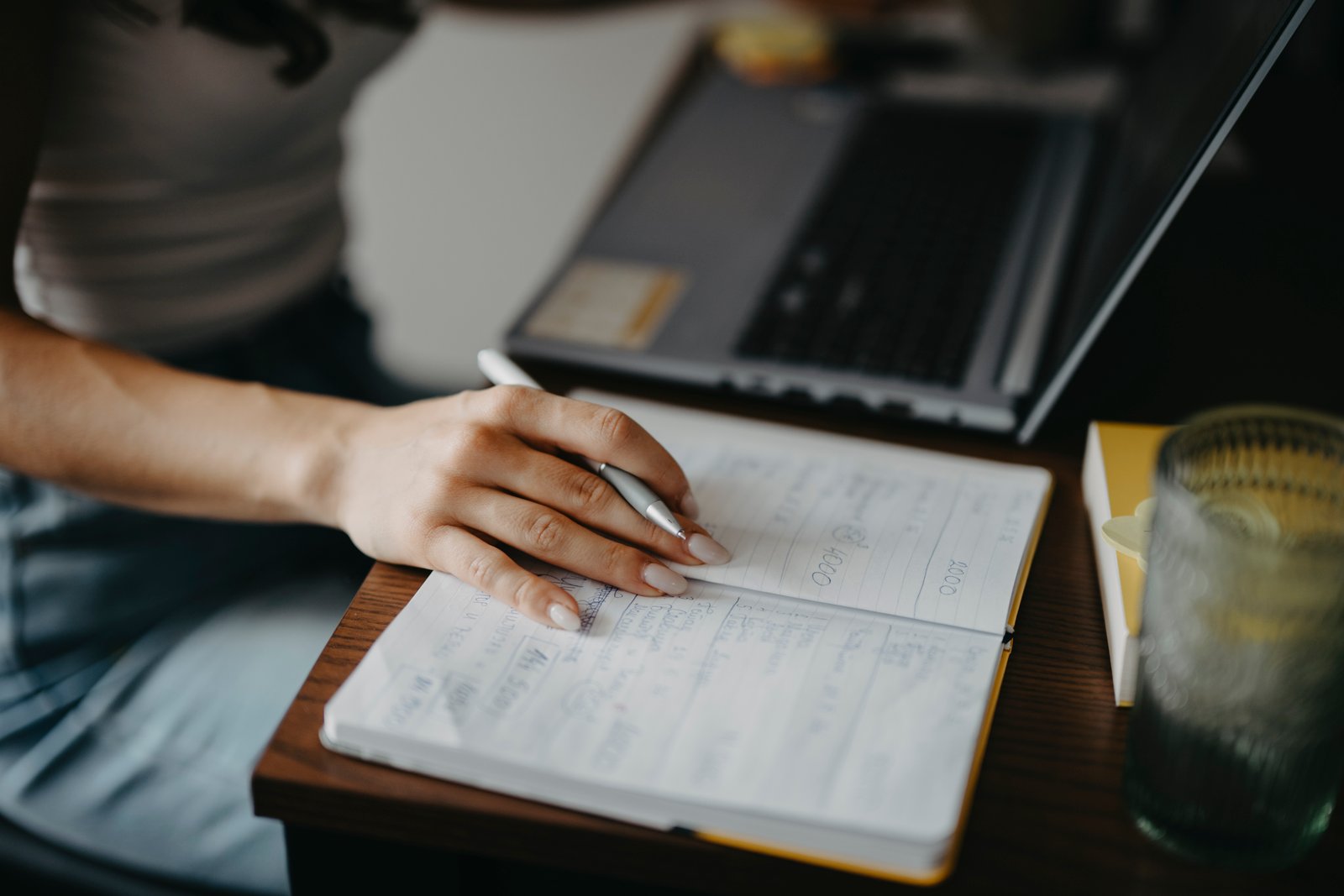 A person writes notes in a lined notebook next to an open laptop on a wooden desk, in the middle of a working session.