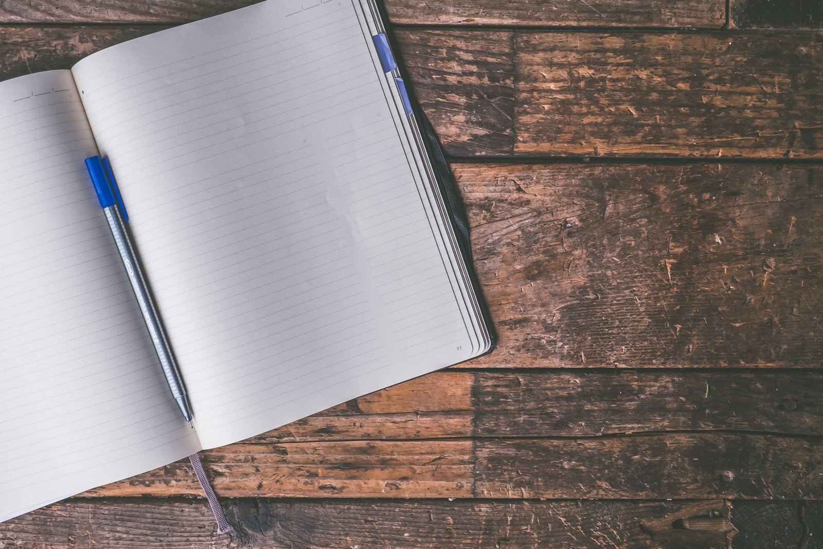 An open notebook with a blue pen resting on a blank lined page, set on a dark wooden desk.