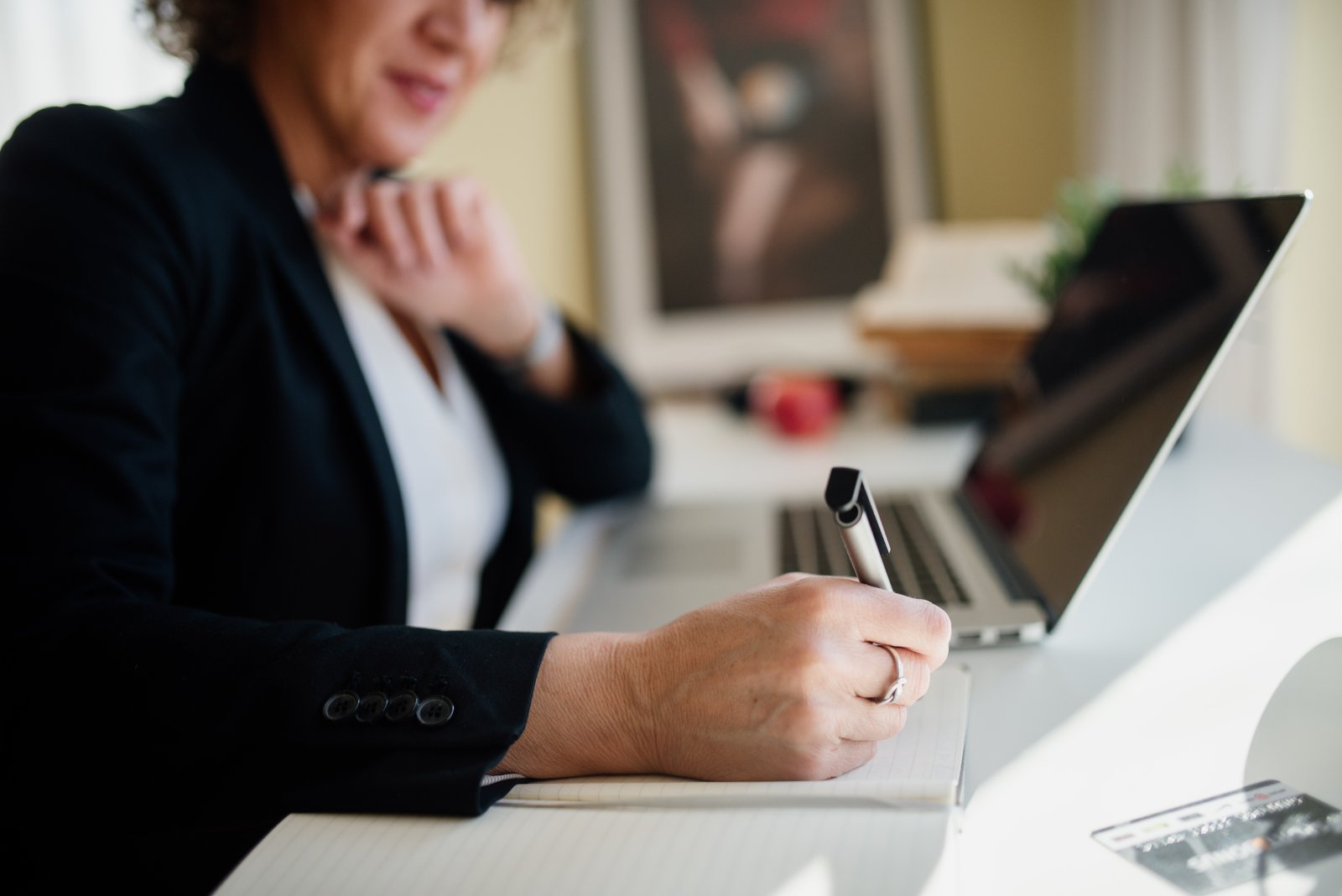 A business professional writing notes in a notebook with a laptop open on the desk beside her