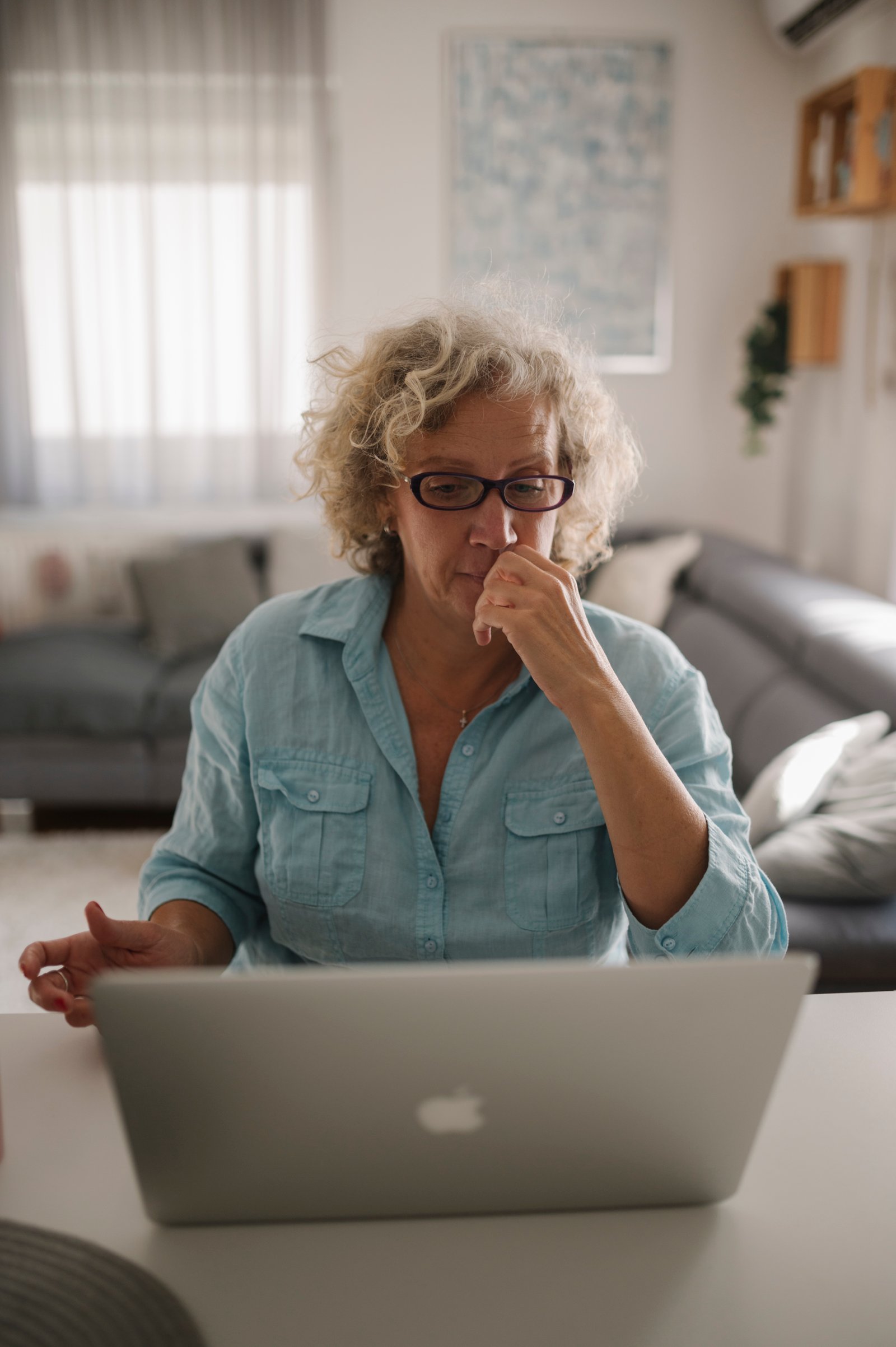 A woman in glasses gazes at a laptop screen with focused, concentrated attention, hand raised to her chin