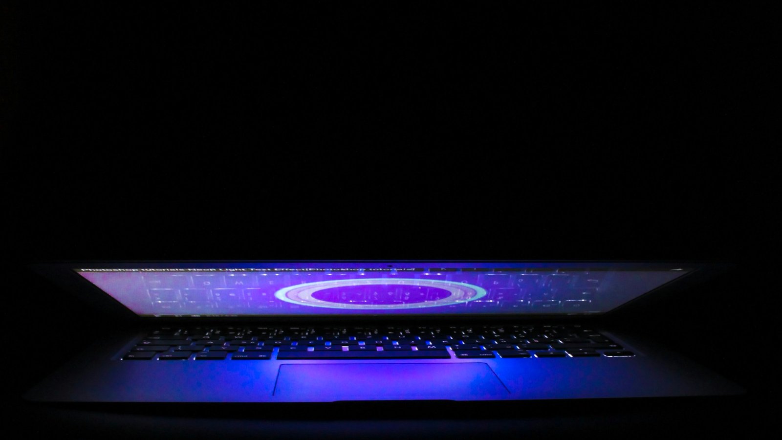 A laptop opened only a sliver in a dark room, electric cyan and violet light spilling from the screen across the keys - the moment before delegating the email you cannot quite write.